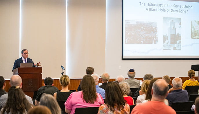 Speaker presents on the Holocaust in the Soviet Union to an attentive audience. A slide titled “A Black Hole or Gray Zone?” is shown on a screen.