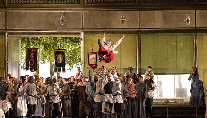 A lively stage scene with performers in traditional attire. A man in red pants is joyfully tossed in the air by a group, surrounded by cheering onlookers and decorations.