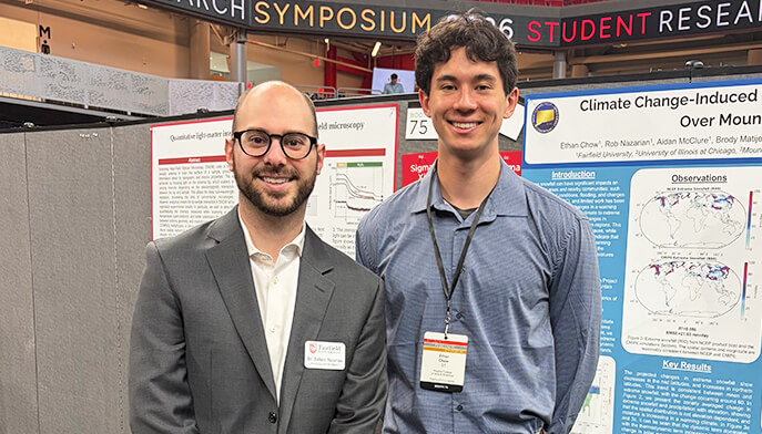 Two men standing in front of research posters at the research symposium. One wears a suit, the other a blue shirt. Both are smiling and wearing name badges.