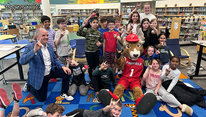 A group of elementary school students poses in a school library with Fairfield University’s mascot, Lucas the Stag, and VP for Athletics Paul Schlickman. The students smile and make "stags up" hand gestures while gathered around the mascot, who is seated on a colorful classroom rug wearing a red Fairfield basketball uniform. Bookshelves and library seating are visible in the background.