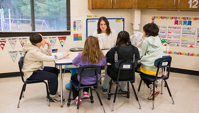 A teacher sits at a round table with five young students in a classroom, engaged in a lesson. Posters and educational materials adorn the walls.