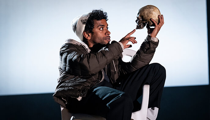 A person in a beanie and jacket sits on a chair, intently holding and pointing at a skull. The setting suggests a dramatic or theatrical scene.