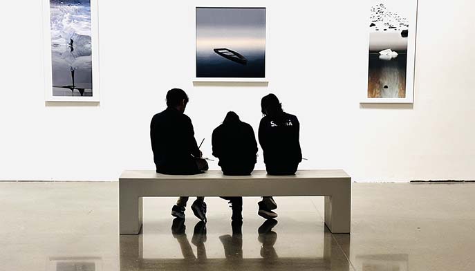 Three people relax on a bench, chatting in front of a wall adorned with various pictures.