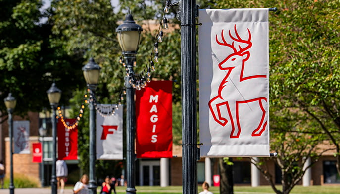 Fairfield University campus walkway with decorative light poles displaying red and white banners featuring the university stag logo and the word "Magis," surrounded by green trees on a sunny day.