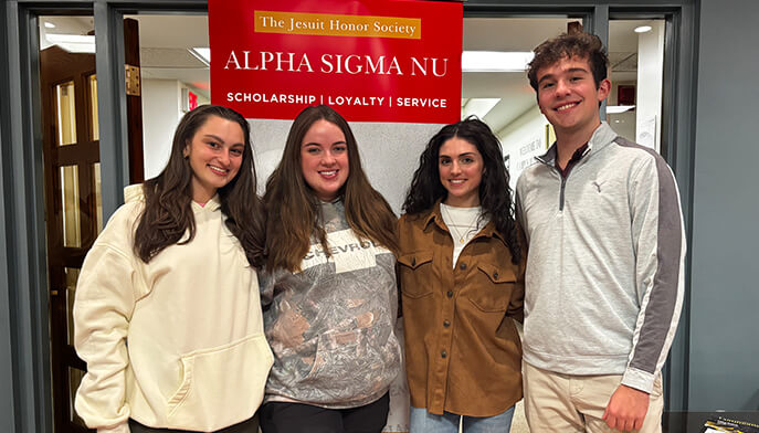 Four smiling students pose beneath an Alpha Sigma Nu banner, celebrating The Jesuit Honor Society’s values of scholarship, loyalty, and service.
