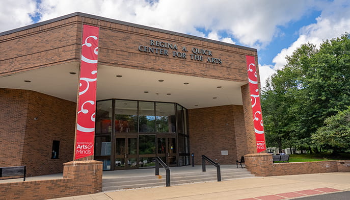 Exterior of Regina A. Quick Center for the Arts, a brick building with red banners, glass doors, trees, and a bright cloudy sky.