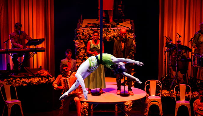 A contortionist arches across a round table during a dramatic cabaret show, surrounded by musicians, singers, red curtains, and flowers.