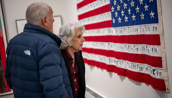 An older couple views a U.S. flag displaying imagery, creating a solemn, reflective memorial scene in an indoor gallery.