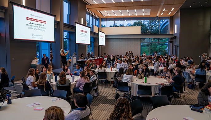 A lively networking event fills a bright conference hall, with attendees seated at round tables, talking while presentation screens hang overhead.