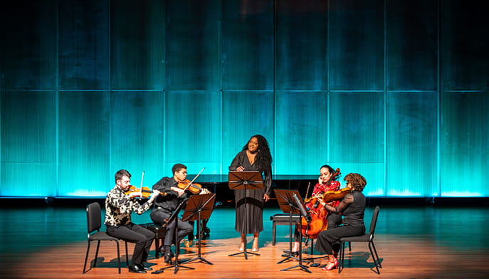 A woman sings at center stage with four seated string musicians playing violin and cello, framed by glowing teal panels and warm wood in an intimate concert setting.