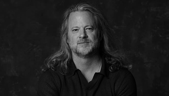 Black-and-white portrait of Nicholas Boggs with long, wavy hair and a short beard, wearing a dark shirt against a textured backdrop; his calm gaze feels thoughtful.