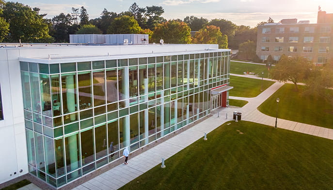 A modern glass-fronted campus building, Fairfield's Barone Campus Center, overlooks manicured lawns and intersecting walkways, with trees and a brick building in warm evening light.