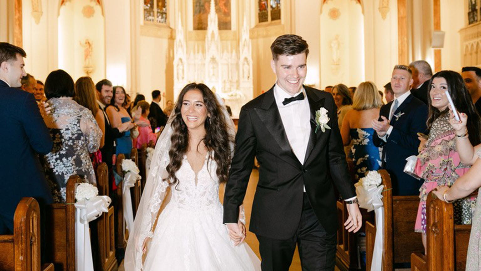 The joyful bride and groom walk down the aisle of an ornate church, smiling broadly. Guests line the pews, clapping and taking photos, capturing the celebratory moment.