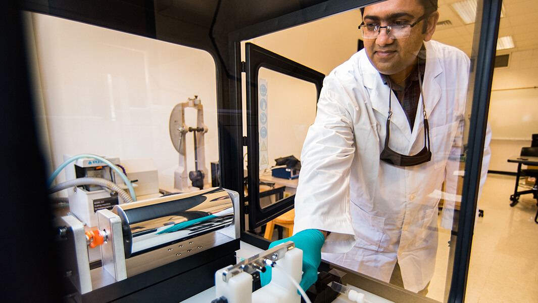 A faculty member in a lab coat and gloves works with a machine inside a lab. The environment is clean and organized, conveying focus and innovation.