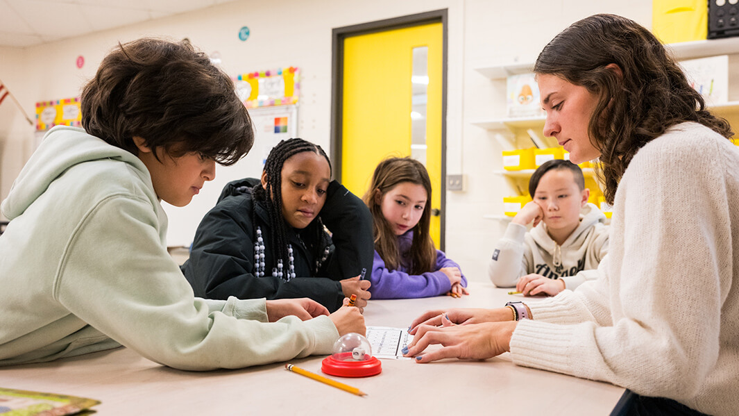 A teacher and four students sit at a table in a classroom, focused on a small robot. The atmosphere is collaborative and engaging, with bright shelves behind.