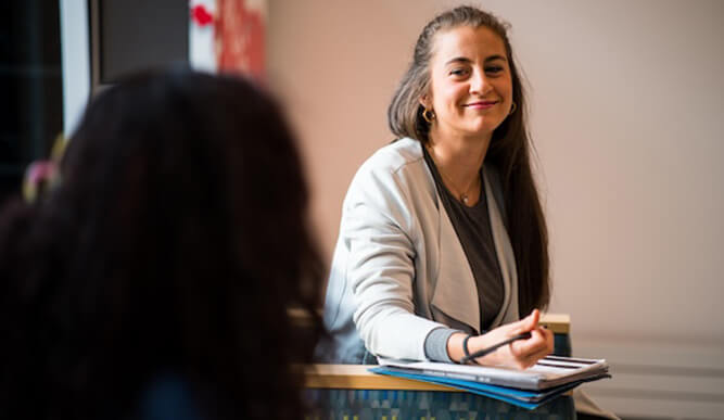 A smiling woman in a light blazer sits at a desk with papers and a pen, speaking with someone in the foreground in a warm, relaxed meeting.
