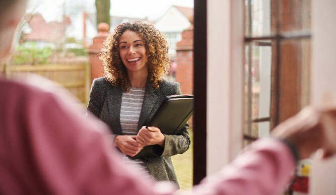 A smiling woman with curly hair holds a folder outside a doorway as someone opens the door, creating a warm, welcoming greeting.