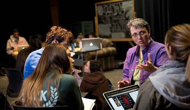 a professor speaks with students in a dimly lit theatre setting.