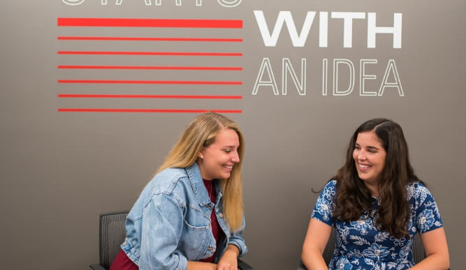 Two dsb students laugh together looking at laptop in front of grey wall.