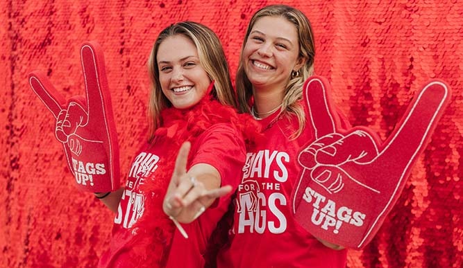 Two students wear red shirts and have foam stag hands in front of a red sequin background.