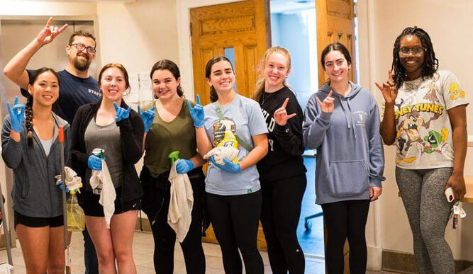 A group of eight smiling people stands indoors, wearing casual clothing and gloves, holding cleaning supplies. They seem cheerful and united, gesturing with peace signs.
