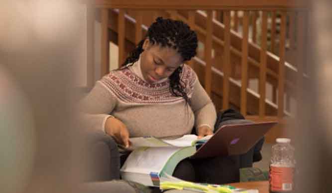 A student reads a textbook while holding a laptop in a library chair.