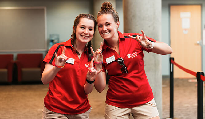 Two female students wearing red shirts and khaki pants stand side by side, smiling for a picture.  