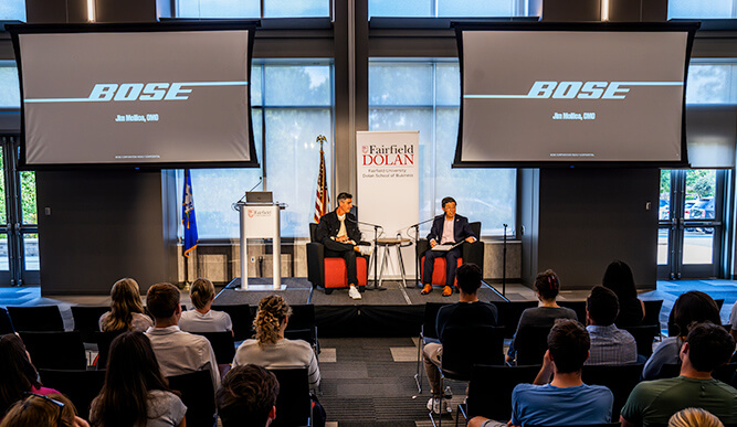 Bose logo displayed on two large screens as two presenters speak on stage to an audience.