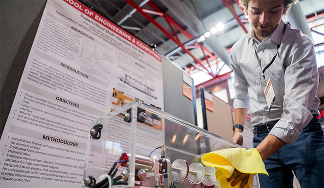 Student cleaning the plexiglass container of a piece of equipment displayed at an annual on-campus research event.