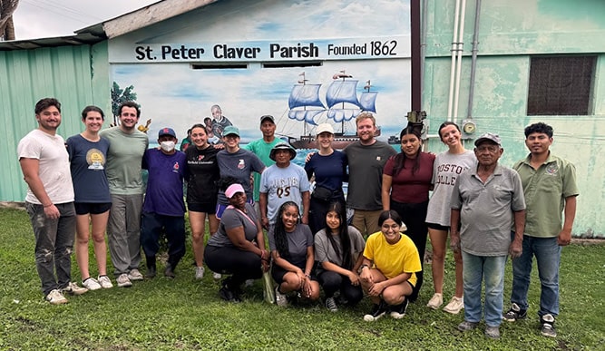 Fairfield students during their immersion trip to Belize, standing in a group in front of a 'St. Claver Parish' mural.