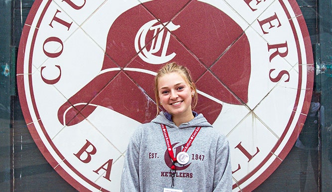 Fairfield Dolan student standing in front of a wall mural of the Cotuit Kettleers Baseball logo while wearing her Cotuit Kettleers sweatshirt.