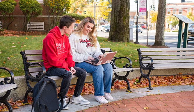 Two Fairfield students sitting on a bench in beautiful downtown Fairfield while completing assignments on a laptop.