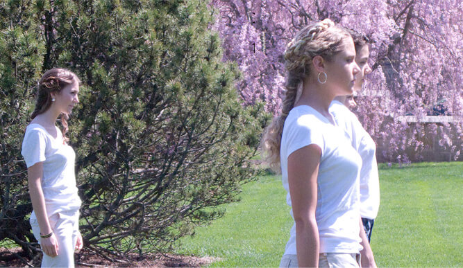A female student in a white shirt stands confidently in a lush green field under a clear blue sky.