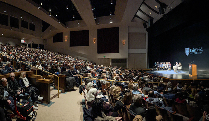 A packed auditorium with people seated, facing a stage with a panel discussion at Fairfield University. The atmosphere is attentive and engaged.