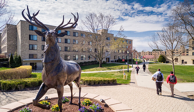 A stag statue stands prominently in front of a brick building on the Fairfield Univesity campus. Students walk on a sunny path, with blue skies overhead.