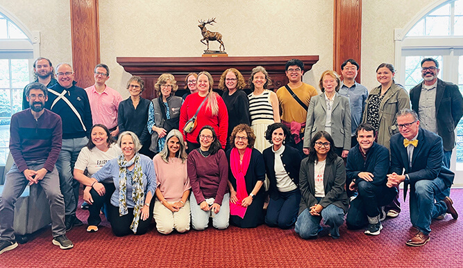 A diverse group of 21 people posing indoors in front of a wooden mantel with a deer statue. The mood is cheerful and casual.