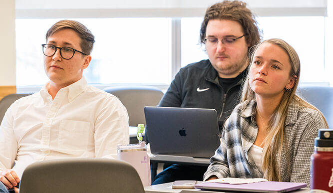 Three students attentively listen in a classroom. Two are seated in the front, one with glasses wearing a white shirt. Another student sits behind, focused on a laptop.