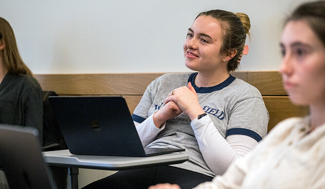  A student sits at a desk with a laptop, listening attentively during a class, with another student in the foreground.