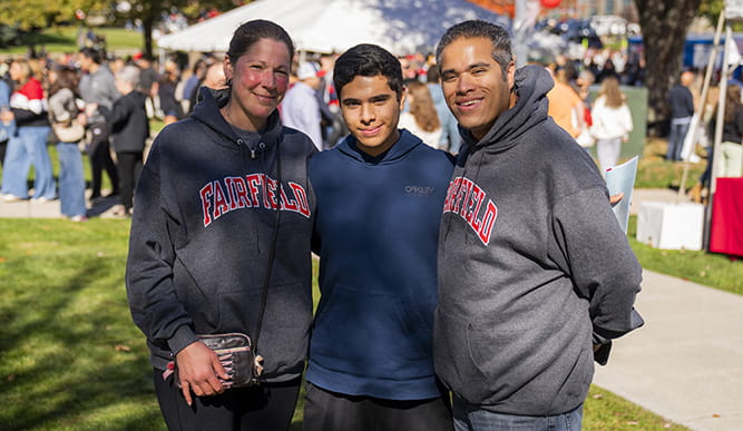 Two adults and a student pose together at an outdoor festival, smiling in Fairfield hoodies, with a sunny campus crowd and tents behind them.