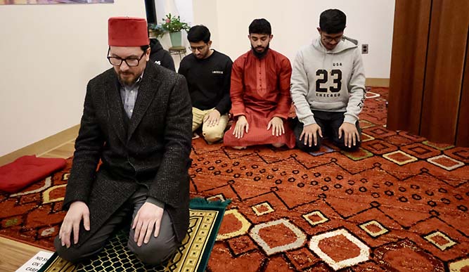 A man wearing a red hat sits on a rug, engaged in a moment of prayer.