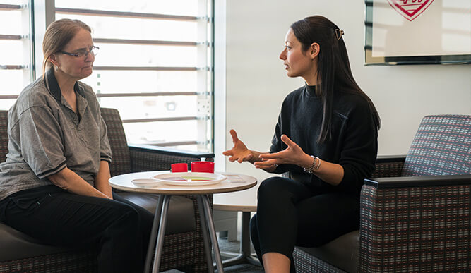 Two women sit across from each other in a bright office area, engaged in conversation; one gestures while the other listens attentively.