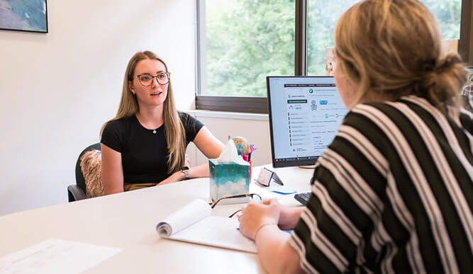 Two women have a conversation across an office desk, with a computer, notebook, and supplies nearby; the scene feels collaborative and professional.