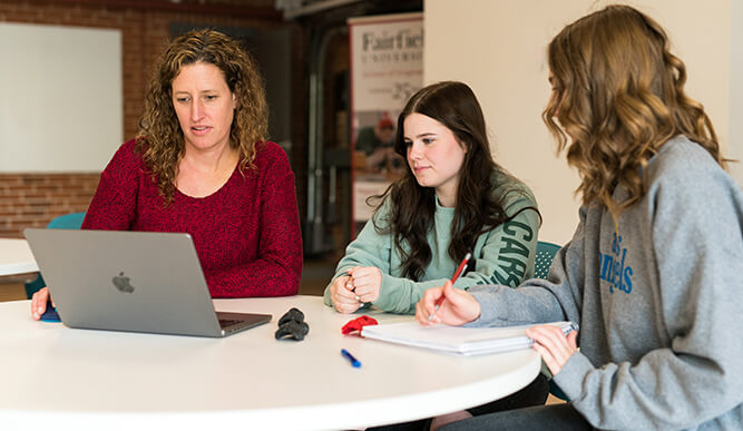 Three women collaborate around a table, reviewing a laptop and papers; focused, engaged discussion in a bright indoor workspace.