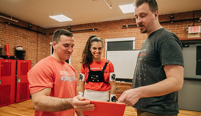 Three people in a brick-walled workshop examine a red tablet, smiling; a woman wears robotic arm components, creating an upbeat training scene.