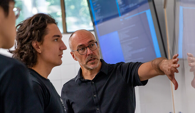 Two colleagues study a computer display as a man gestures toward the screen, explaining technical information in a collaborative setting.