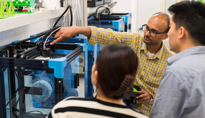 Engineer demonstrates a 3D printer in a laboratory to three colleagues, pointing at its components during a focused training session.