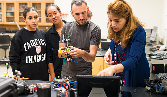 Focused students and instructors collaborate in an electronics lab, examining a colorful drone circuit amid tools and equipment.