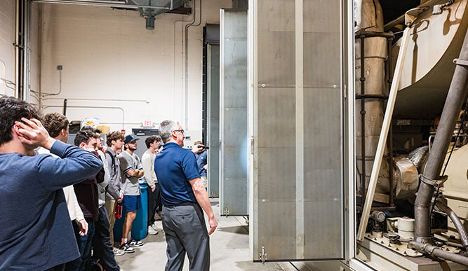 A group tours an industrial facility, watching machinery behind a large open metal door; the scene feels curious, educational, and hands-on.