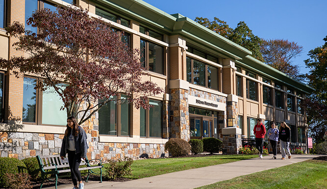 DiMenna-Nyselius Library with stone facade, leafy trees, benches, and students walking along a sunny sidewalk under blue sky.
