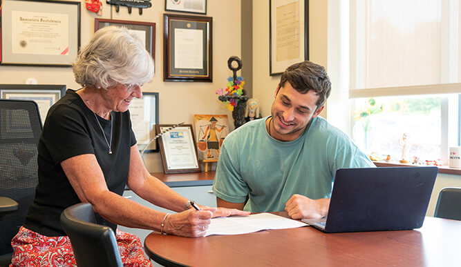 Older woman and young man review paperwork together at an office table beside an open laptop, conveying a warm, collaborative consultation.